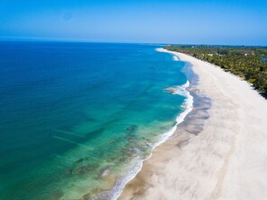 An aerial view of a beach with clear turquoise waters, a sandy shoreline, and a line of trees and buildings along the coast.