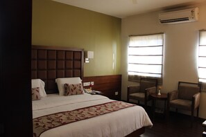 A hotel room with a large bed, a wooden headboard, a bedside table with a lamp, two chairs, a small table with a vase, and an air conditioning unit. - Stay Inn (Hyderabad)