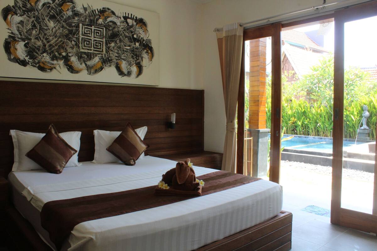 A hotel room with a large bed, a headboard, a wall-mounted artwork, and a sliding glass door leading to an outdoor area.