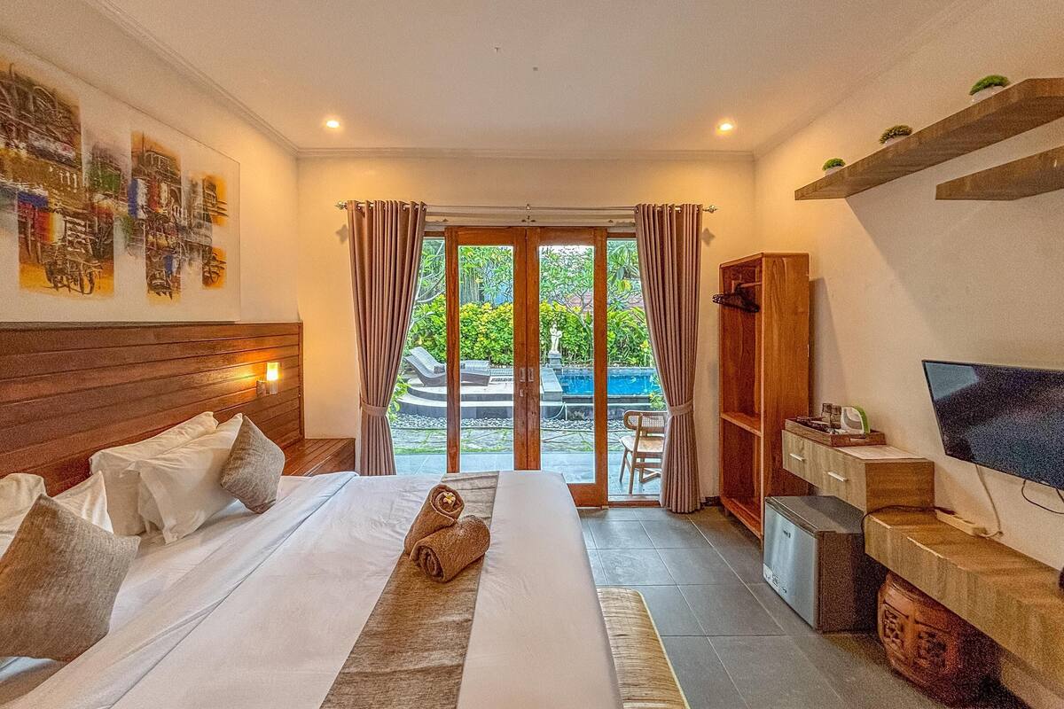 A hotel room with a bed, a TV, a desk, and a view of a pool and garden.