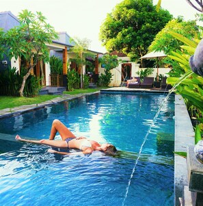 A swimming pool with a person lying on the edge, surrounded by lush greenery and a building in the background.