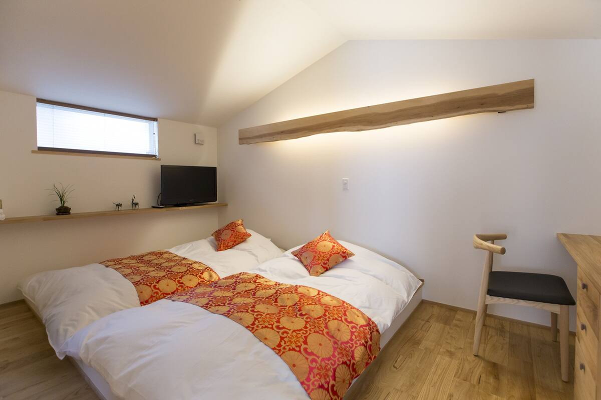 A bedroom with two beds, a TV mounted on the wall, a wooden bench, and a shelf with a plant.