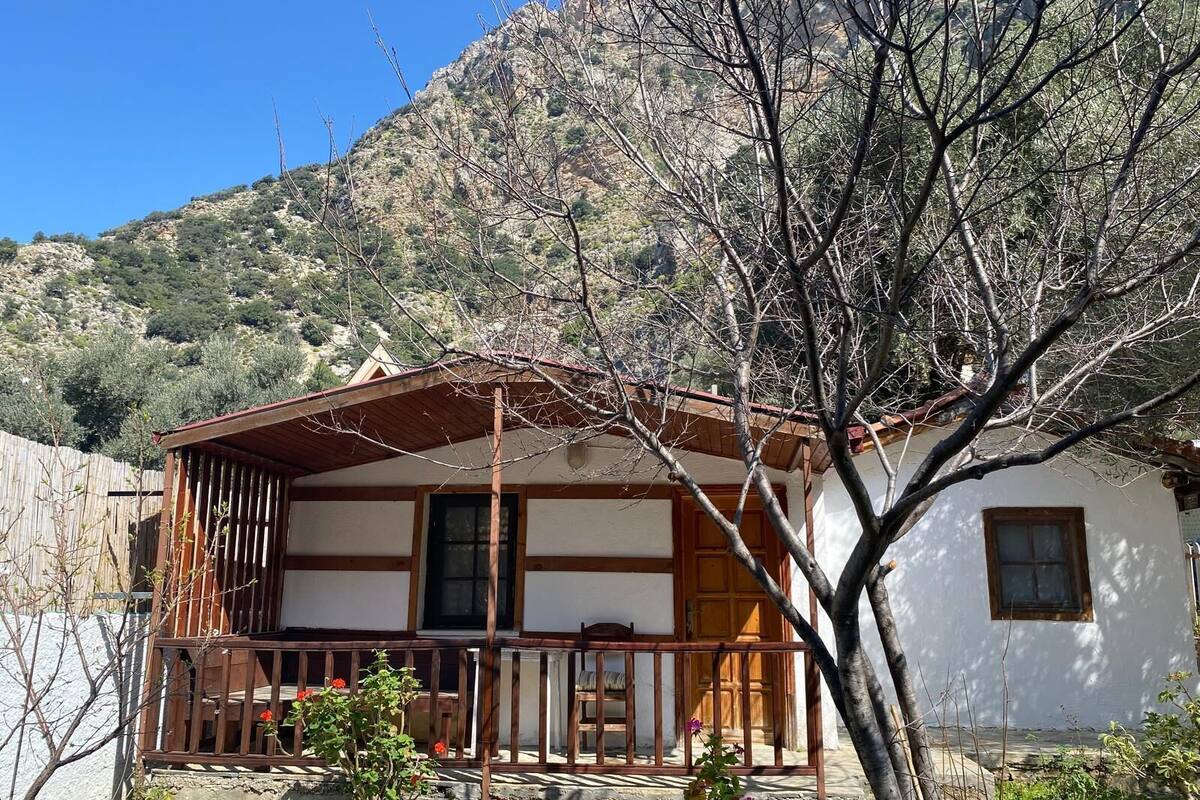 A traditional wooden house with a balcony, surrounded by a stone wall and a mountain backdrop.