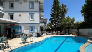 A swimming pool with lounge chairs and tables, surrounded by a building with multiple balconies and a clear blue sky. - Gozde Hotel (Serik)