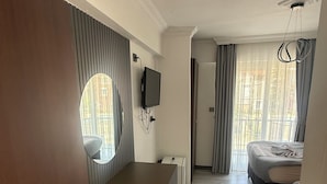 A hotel room with a wooden desk, a television mounted on the wall, a bed with a grey bedspread, and a large window with curtains. - Gozde Hotel (Serik)