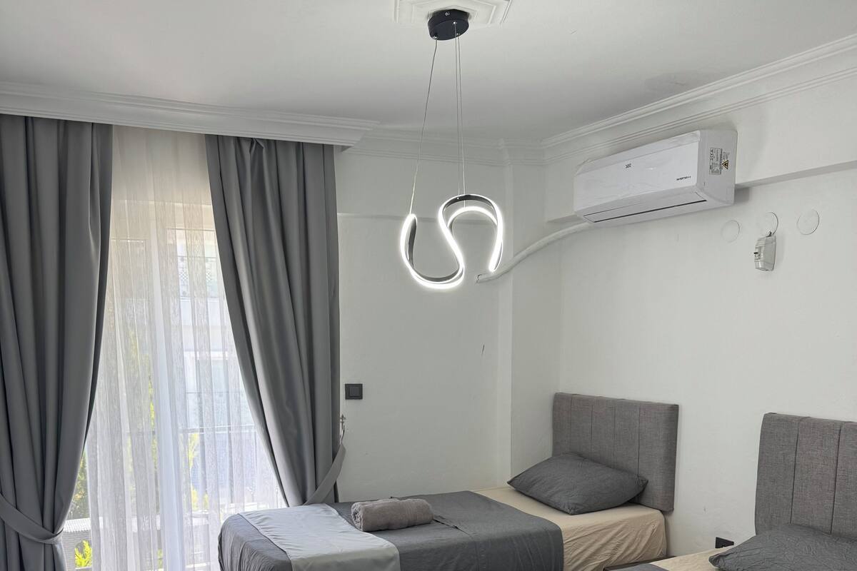 A modern hotel room with two beds, a ceiling light fixture, an air conditioning unit, and a view through the window.