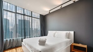Camera da letto moderna con un letto grande, un comodino in legno, tende trasparenti e vista sulla città.