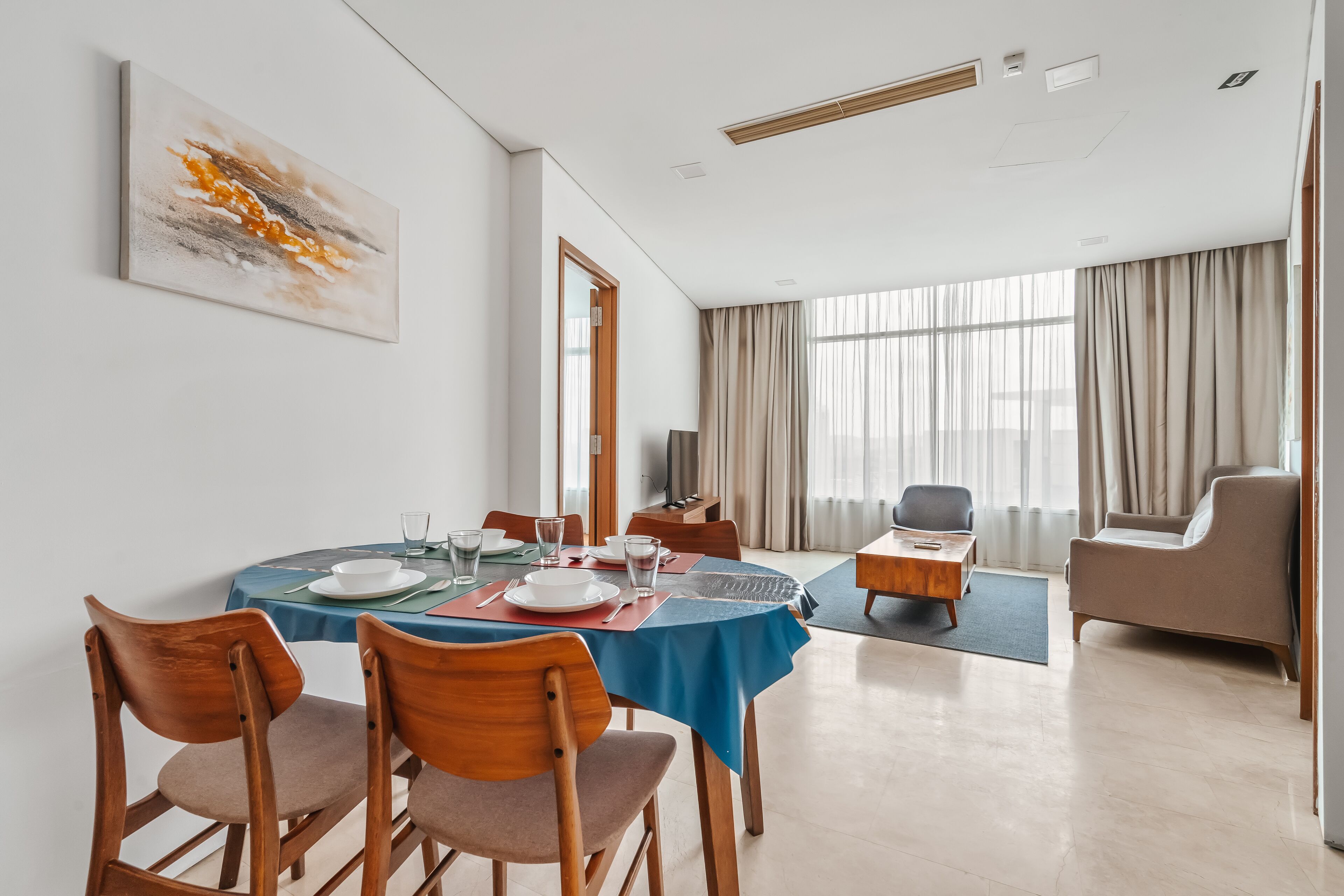 Ein moderner Essbereich mit einem runden Tisch, Holzstühlen und einer blauen Tischdecke. Es gibt ein großes Fenster mit Vorhängen, eine Couch und einen Fernseher.