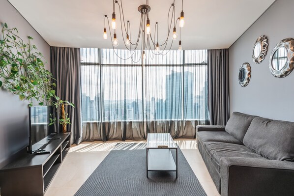 Una sala de estar moderna con un sofá gris, una mesa de centro, un televisor sobre un mueble, una araña y cortinas translúcidas, con vistas a la ciudad.