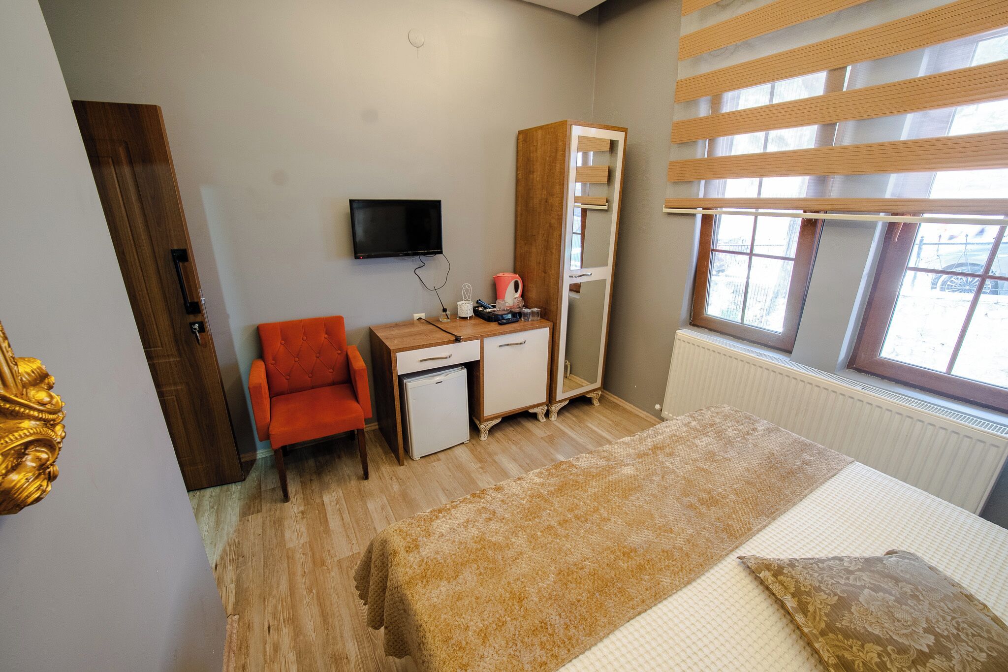 A bedroom with a bed, a desk with a chair, a wardrobe, a television, and a window with blinds.