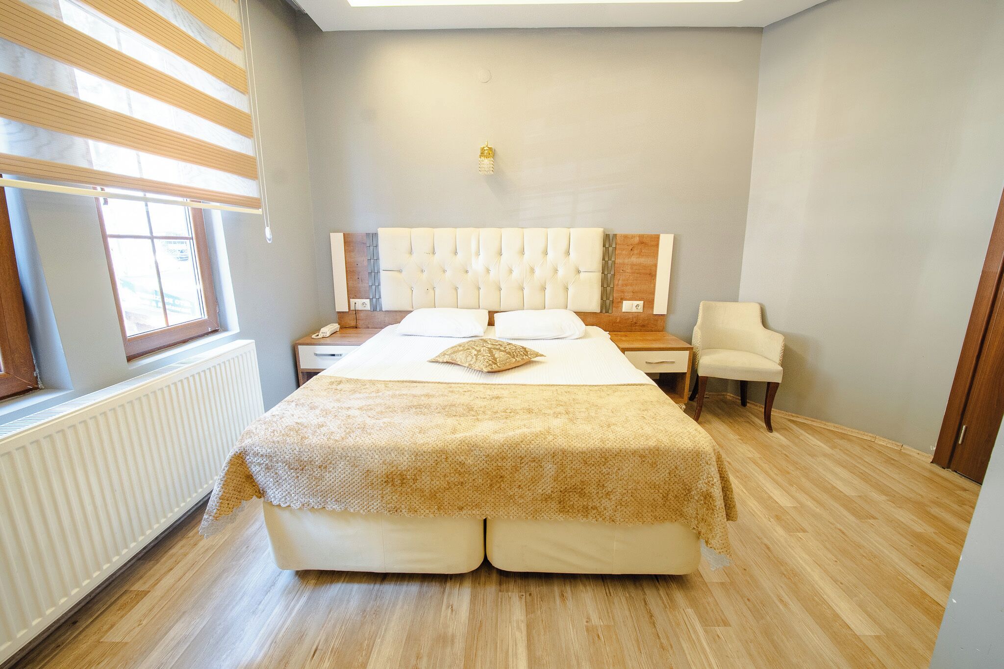 A bedroom with a large bed, a bedside table, a chair, a window with blinds, and a radiator.