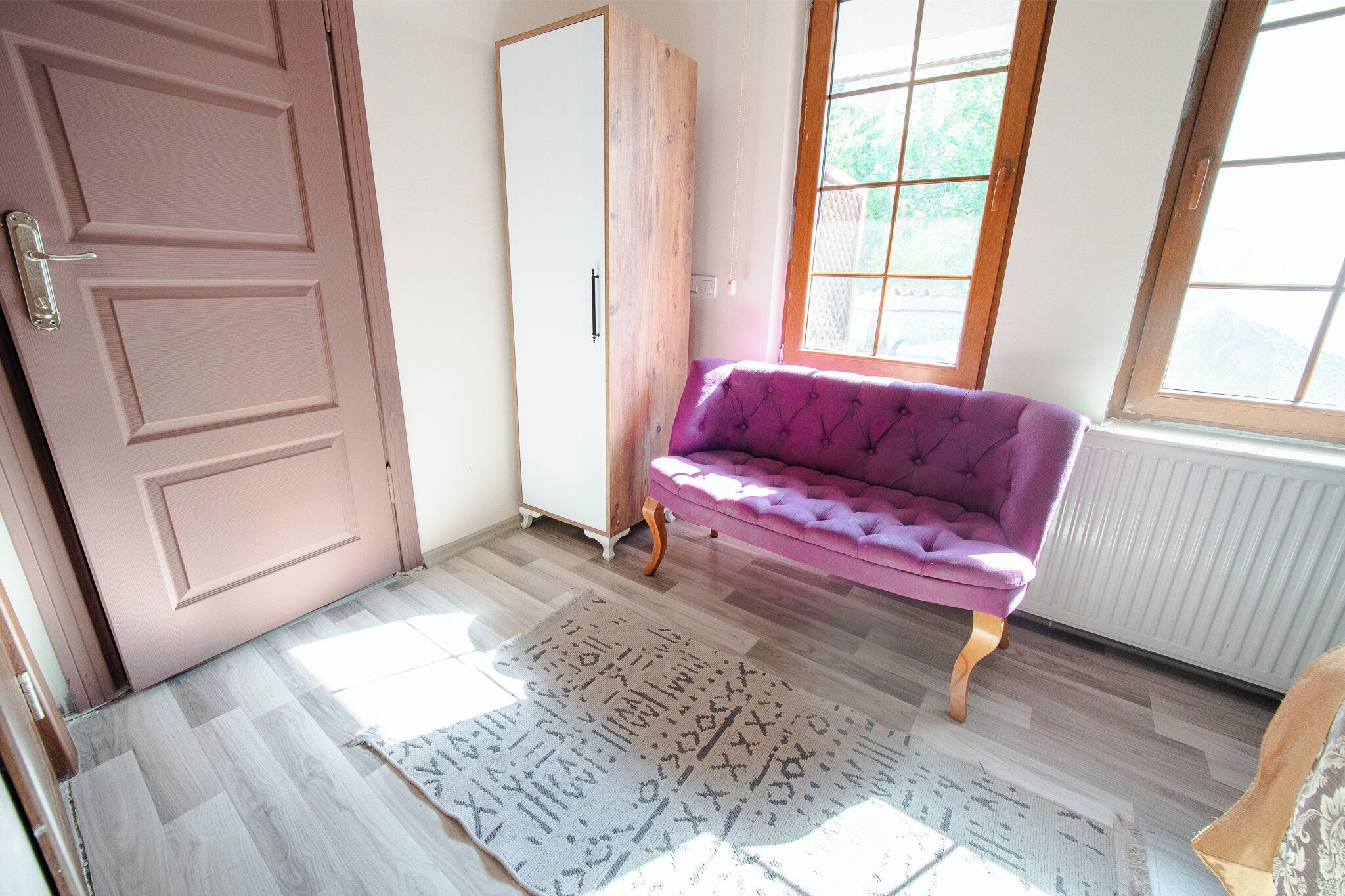 A room with a wooden floor, a pink door, a purple sofa, and a window with a view of greenery.