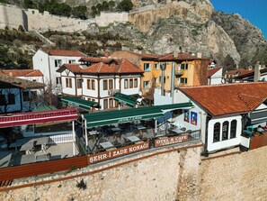 A traditional building with a stone facade, a sign for 'SEHR-I ZADE KONAĞI' indicating a cafe, and a view of a mountainous landscape in the background.