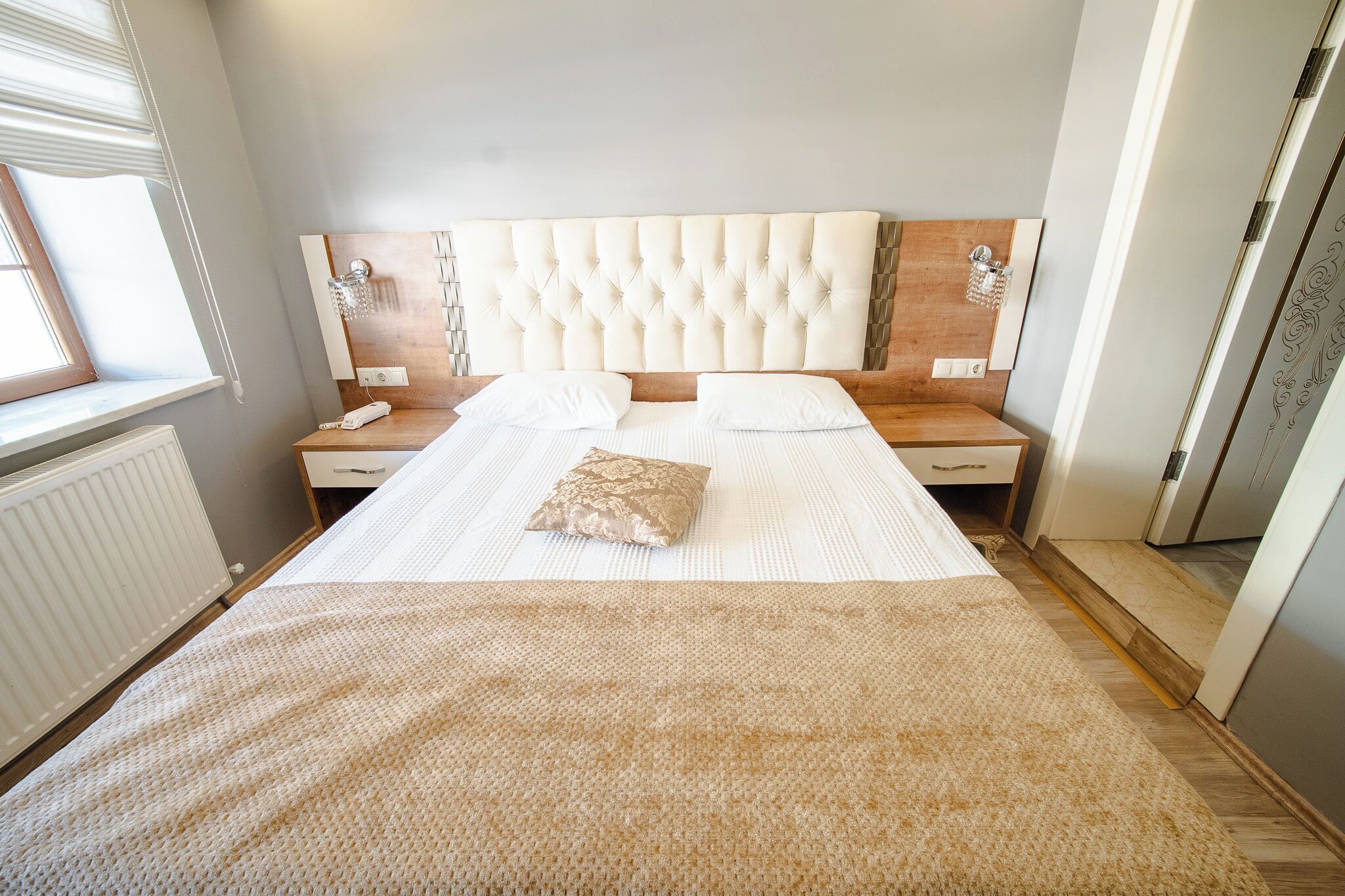 Une chambre à coucher avec un grand lit, une tête de lit en bois, deux tables de chevet, une commode, une fenêtre et un radiateur.