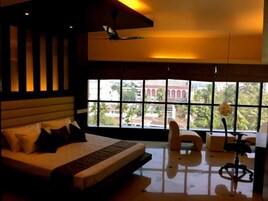 A bedroom with a bed, a desk, and a chair. There is a large window with a view of buildings and trees.