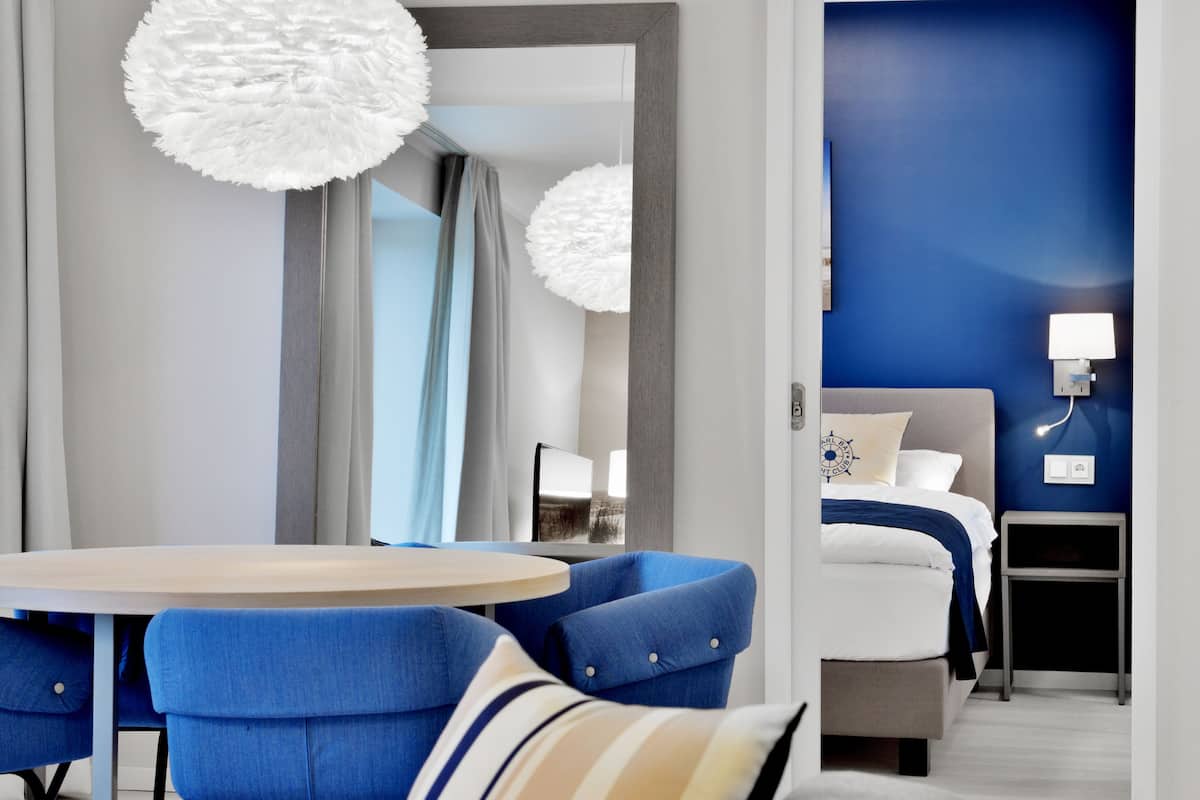 Ein modernes Hotelzimmer mit einem runden Esstisch, blauen Polsterstühlen, einem großen Spiegel, einem Bett mit blauer Akzentwand und einem Nachttisch mit Lampe.