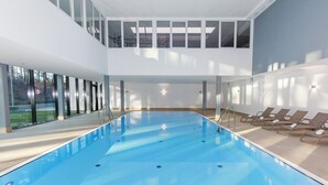 Une piscine intérieure moderne, avec une eau d’un bleu cristallin, des chaises longues et de grandes fenêtres offrant une vue sur l’extérieur.
