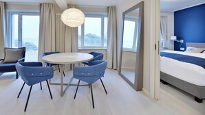 Une chambre d’hôtel moderne avec un grand lit, une table de salle à manger ronde entourée de chaises, un canapé et une vue sur l’extérieur grâce à de grandes fenêtres.
