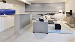 Un salon moderne doté d’un grand téléviseur à écran plat, d’un canapé gris et offrant une vue sur la plage grâce à une grande fenêtre.