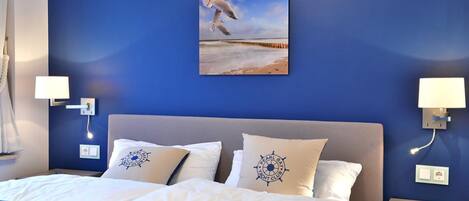 Una habitación de hotel con una cama grande, dos almohadas con un logo, un cuadro de una gaviota y una mesita de noche con una lámpara.