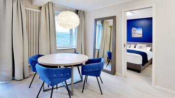 A modern hotel room with a round dining table, blue chairs, a large window with curtains, a bed with a blue headboard, and a painting on the wall.
