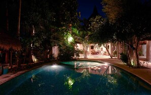 A nighttime pool area with a clear pool, surrounded by tropical vegetation and thatched-roof structures.