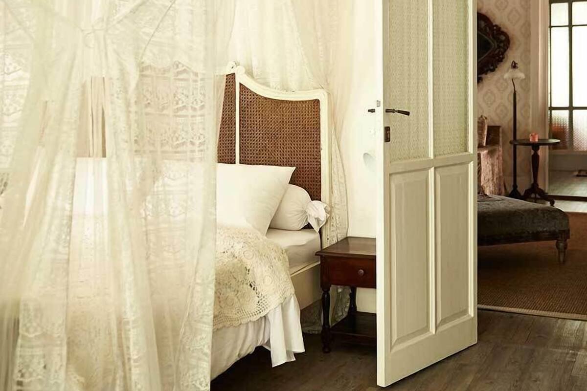 A bedroom with a canopy bed, a wooden floor, and a door leading to another room.