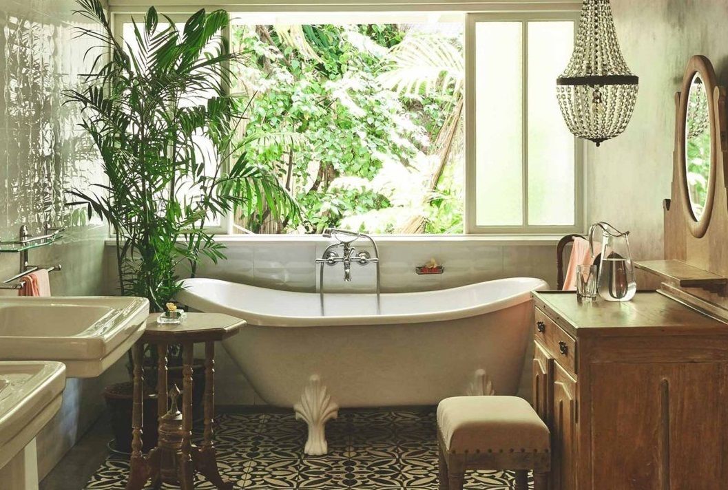 A bathroom with a freestanding bathtub, a window with a view of greenery, a chandelier, and a wooden vanity.