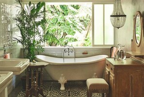 Een badkamer met een vrijstaand bad, een raam met uitzicht op groen, een kroonluchter en een houten wastafelmeubel.