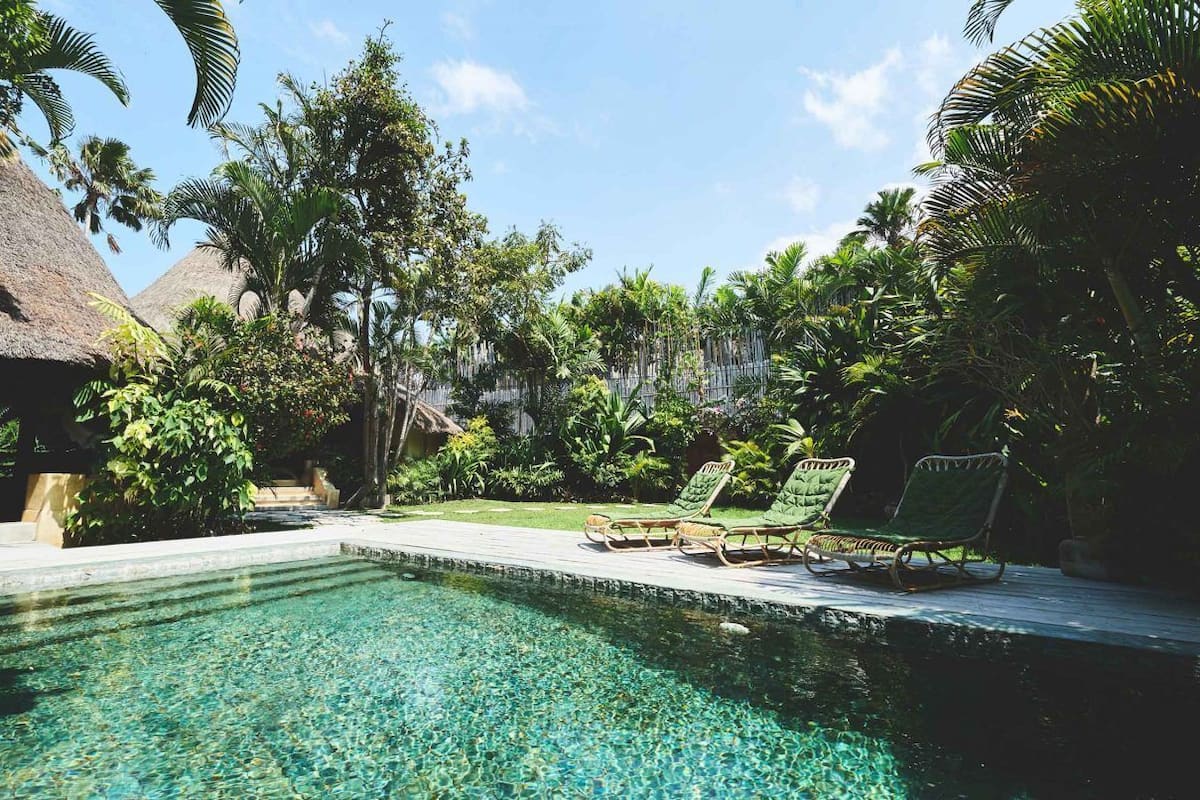 A pool with clear water, surrounded by lush greenery and thatched-roof structures.
