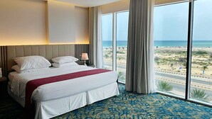 A hotel room with a large bed, a nightstand, a lamp, and a view of the beach. - Surgrand Hotel (Sur)