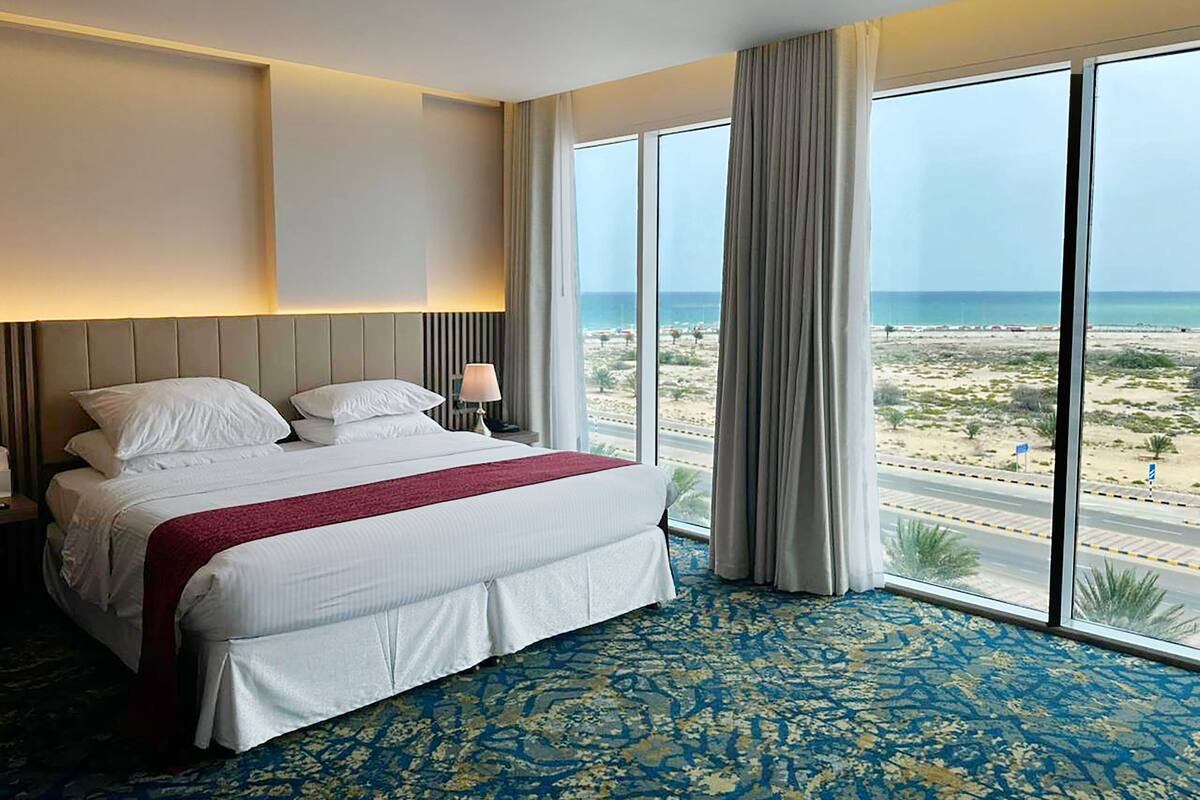 A hotel room with a large bed, a nightstand, a lamp, and a view of the beach.