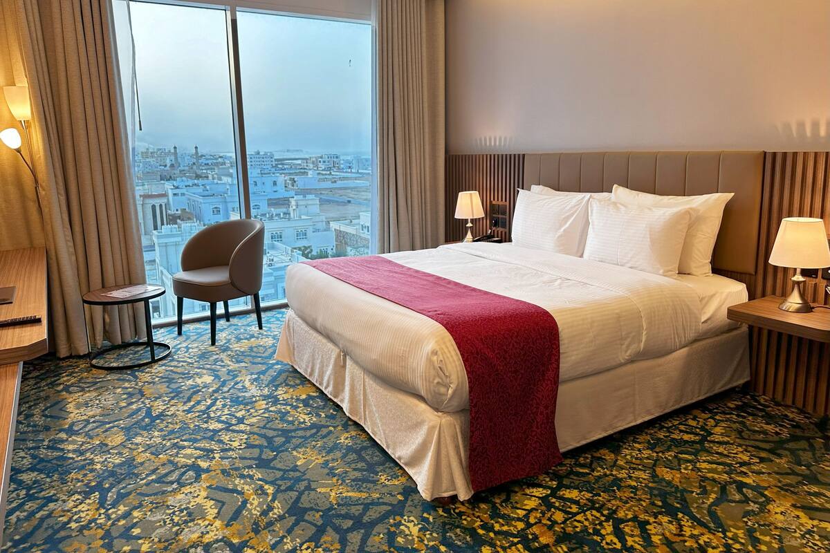 A hotel room with a large bed, two bedside tables, a chair, and a view of a cityscape through large windows.