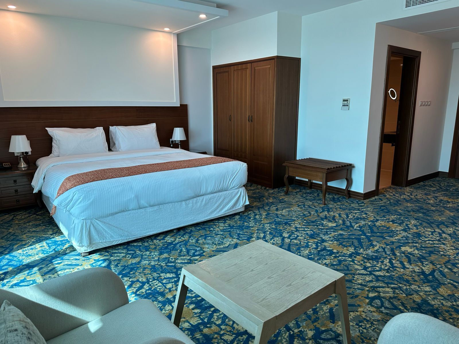 A hotel room with a large bed, a wooden wardrobe, a small table, and a seating area with chairs.