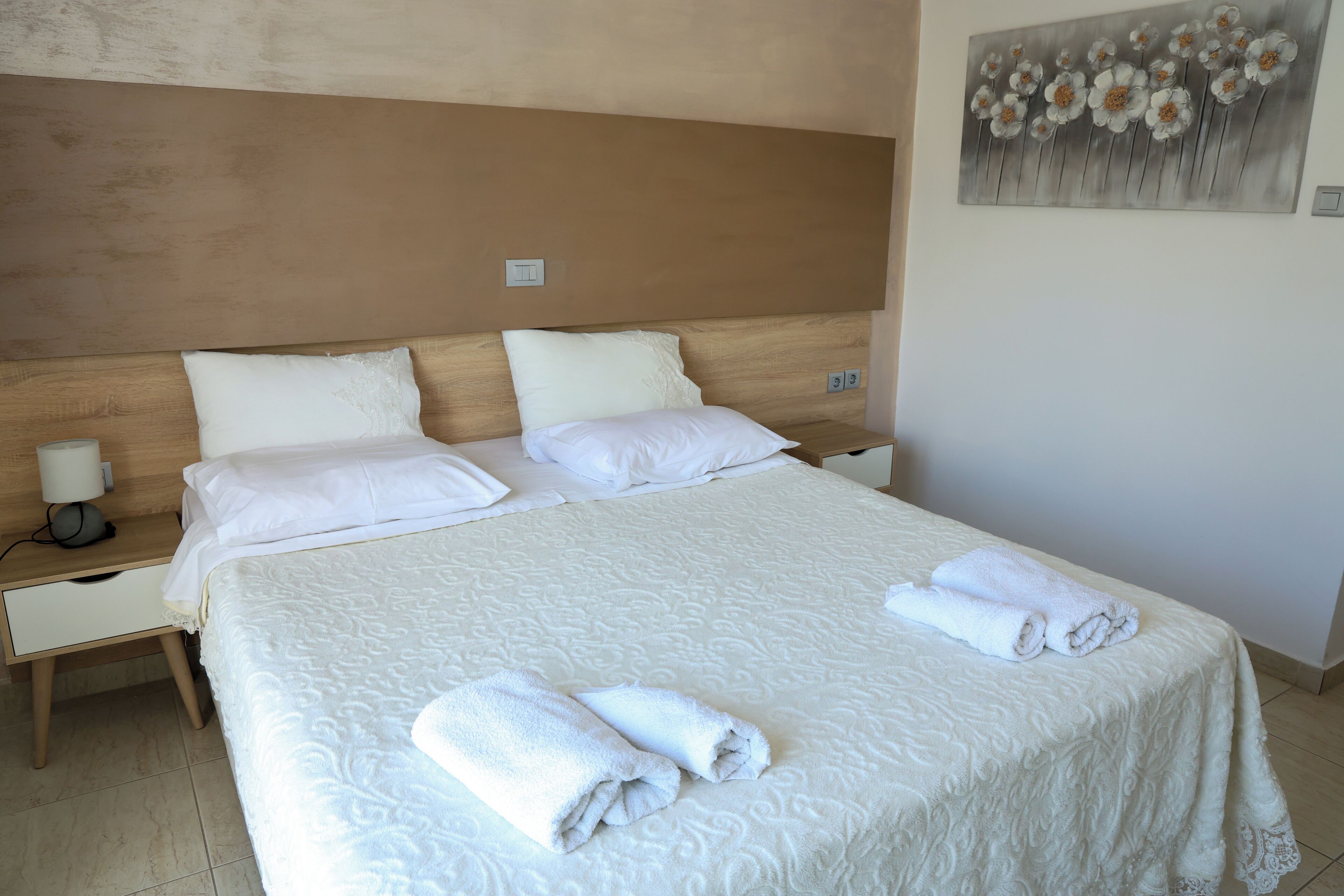 A neatly made bed with white linens and rolled towels, a wooden headboard, and a painting on the wall.