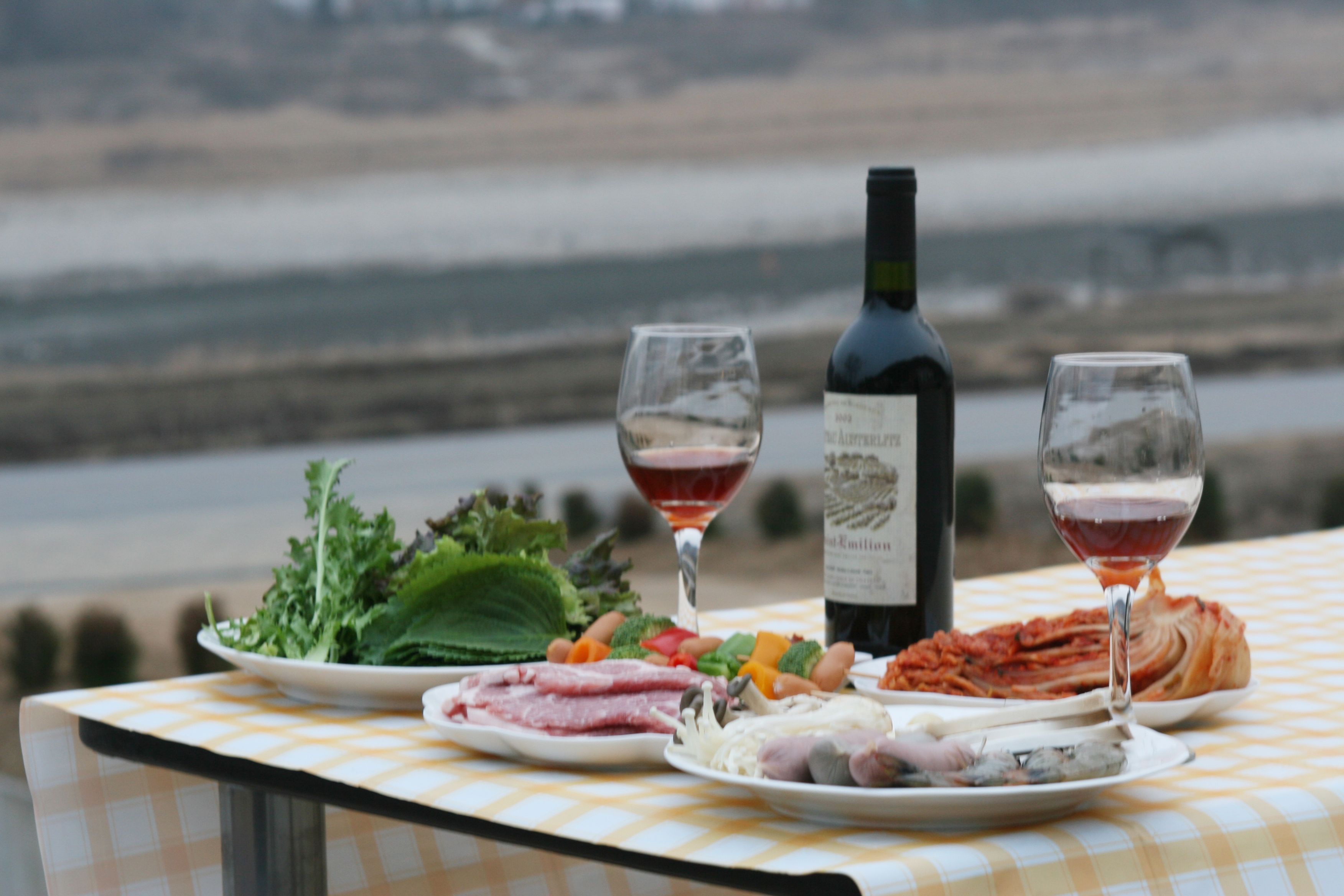 A table with a checkered tablecloth, two wine glasses, a bottle of wine, and plates of food including vegetables, meat, and cheese.