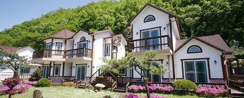 Una casa de dos pisos con un balcón, un porche delantero y un pequeño jardín con flores rosas.