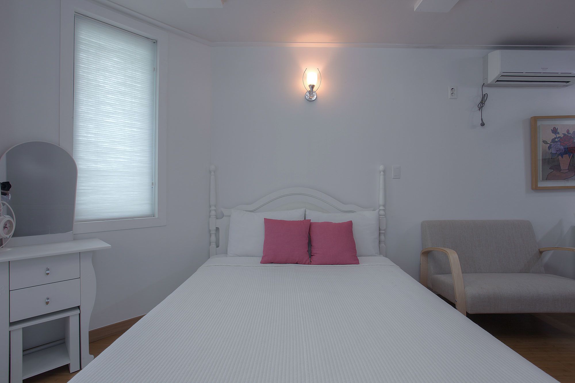 Un dormitorio con una cama blanca, una mesita de noche, una silla y una lámpara fijada a la pared.