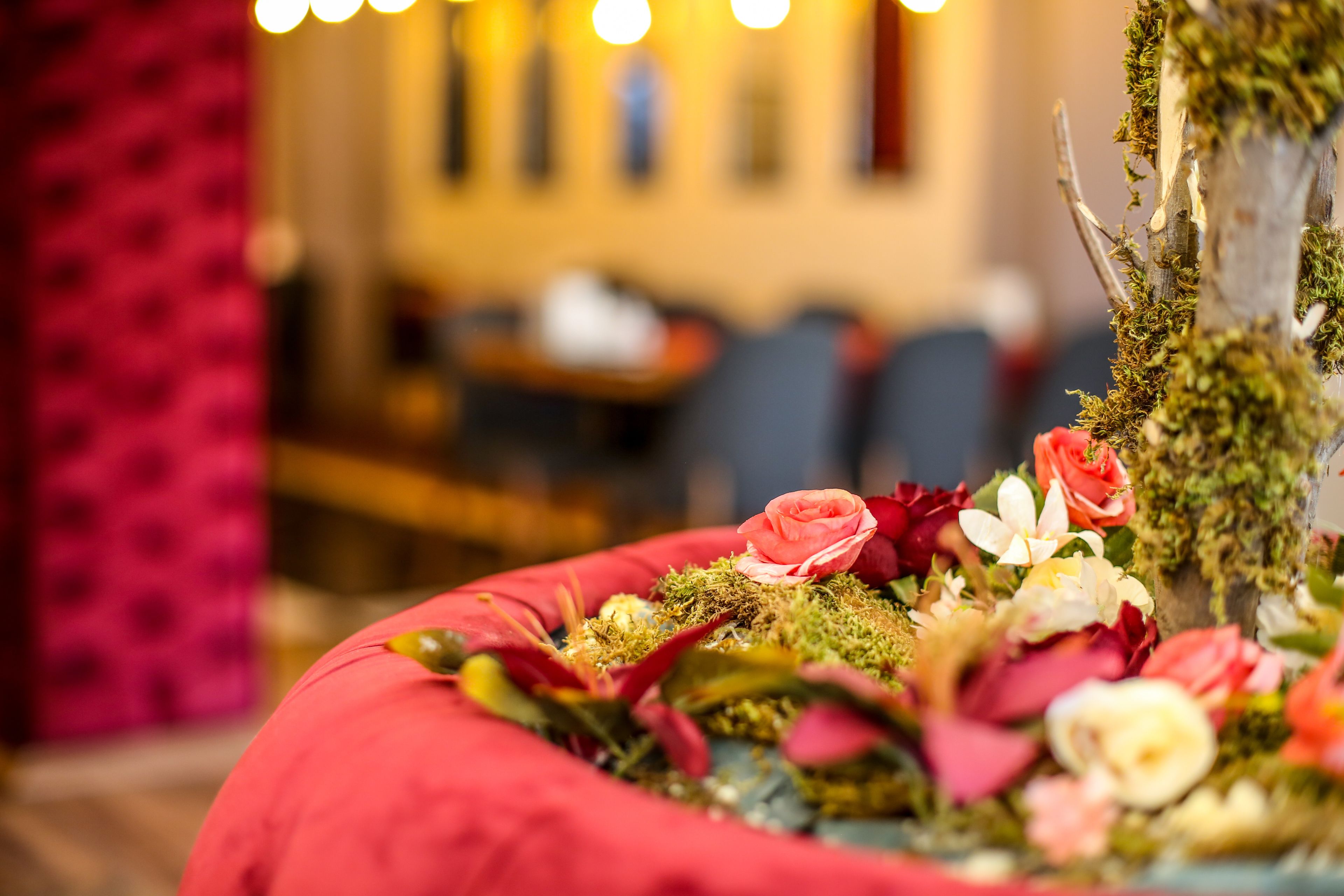 Nahaufnahme einer Blumenkomposition mit verschiedenen Blumen und Grünpflanzen vor unscharfem Hintergrund eines Raumes mit roter Wand und Tisch.