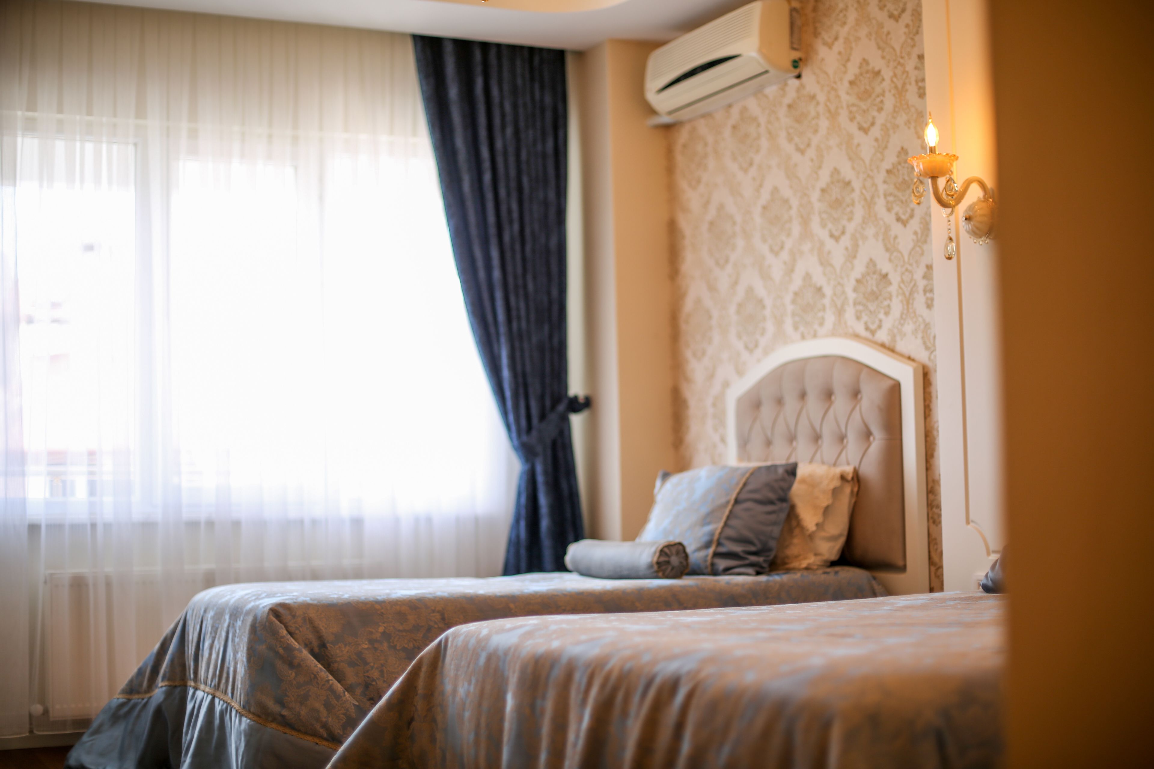 A hotel room with two beds, a large window with sheer curtains, an air conditioning unit, and a wall-mounted light fixture.