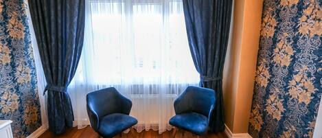 Una habitación con piso de madera, dos sillones azules, una cama con colcha estampada y una ventana con cortinas traslúcidas.