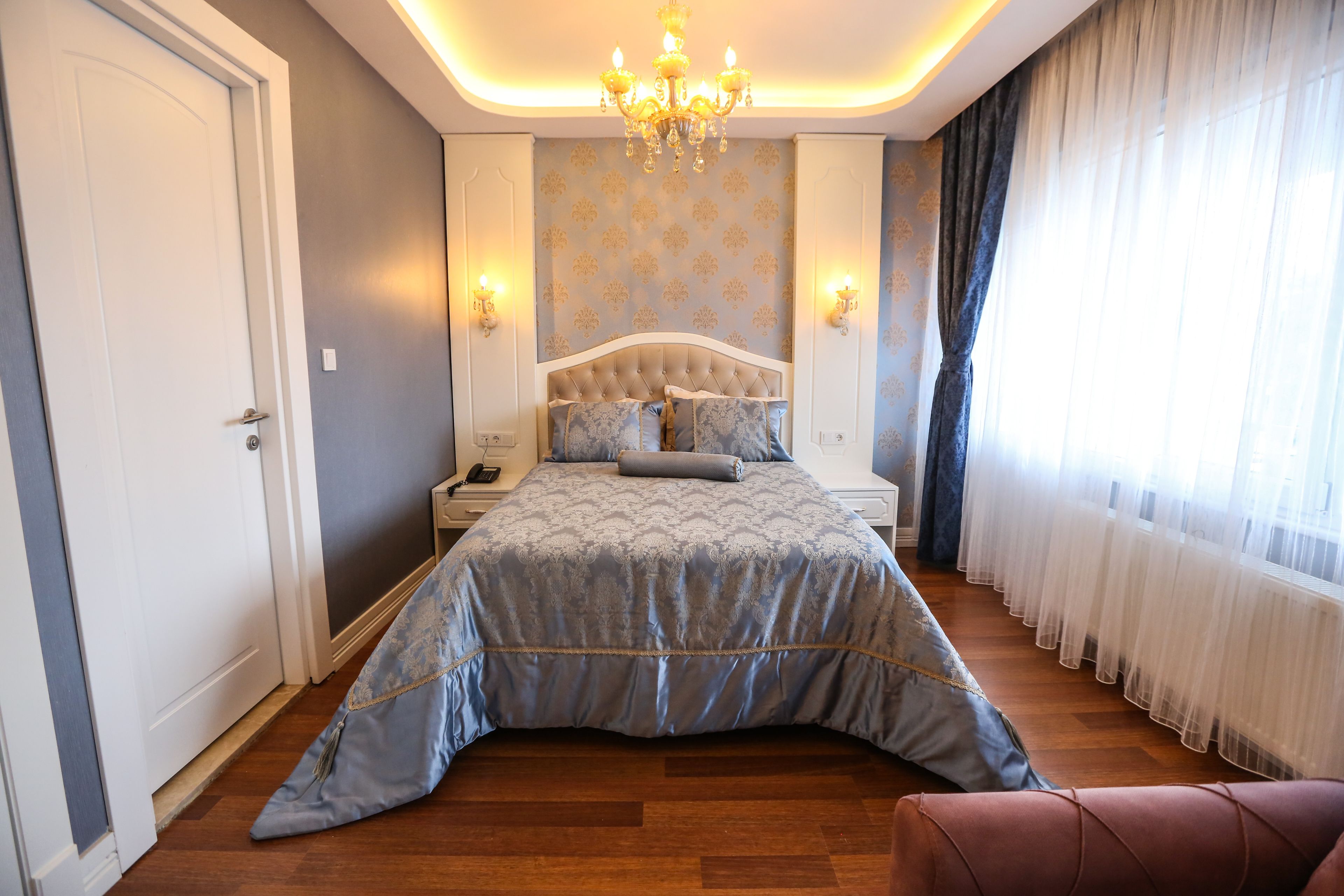 A bedroom with a large bed, a chandelier, a nightstand, and a window with curtains.