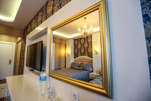 Ein Hotelzimmer mit einem großen Spiegel, einem Fernseher, einem Bett mit einem gemusterten Überwurf, einer Kristallleuchter und einer Wasserflasche auf der Ablagefläche.