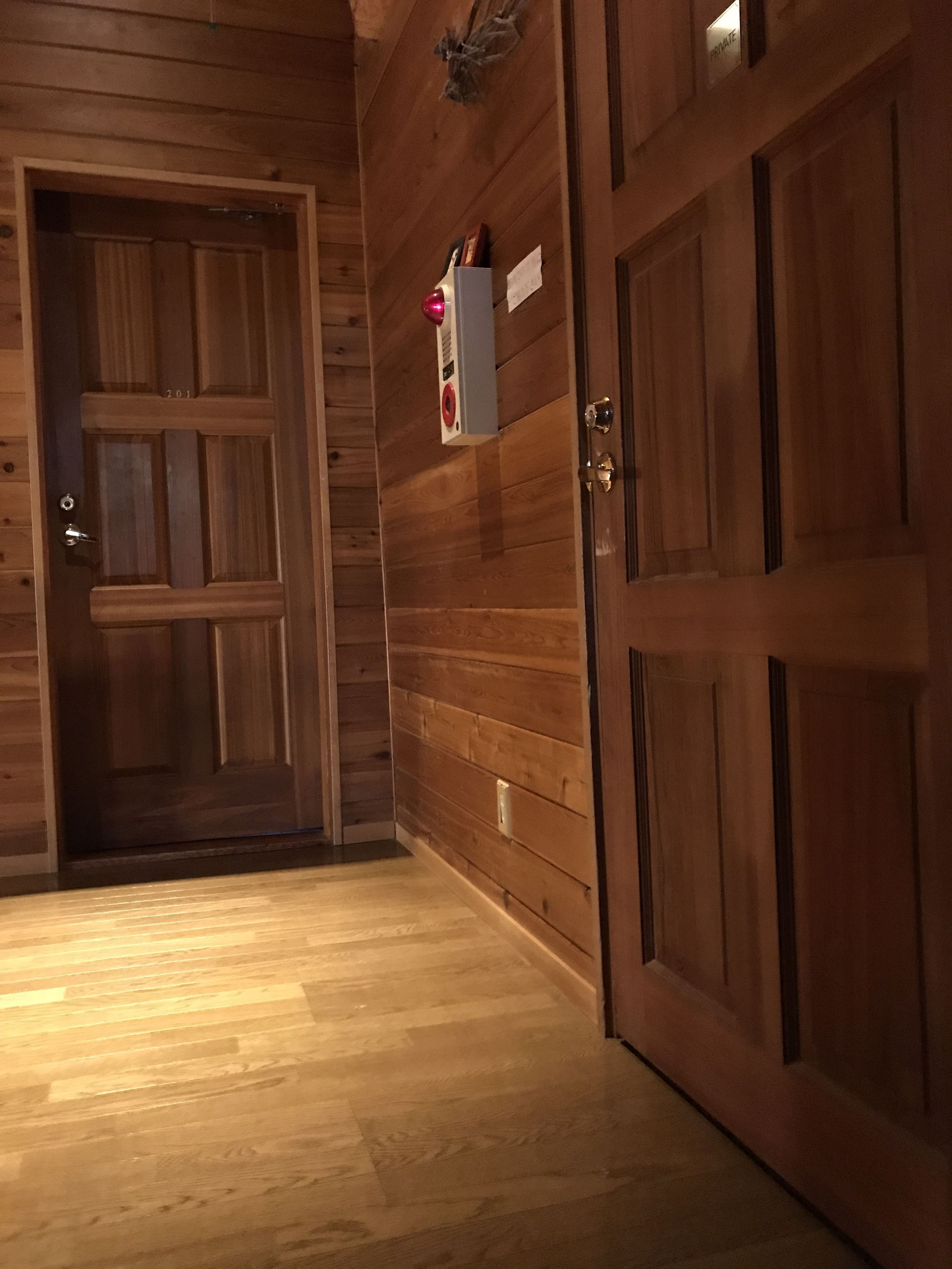 A wooden hallway with a closed door, a wall-mounted fire alarm, and a light switch.