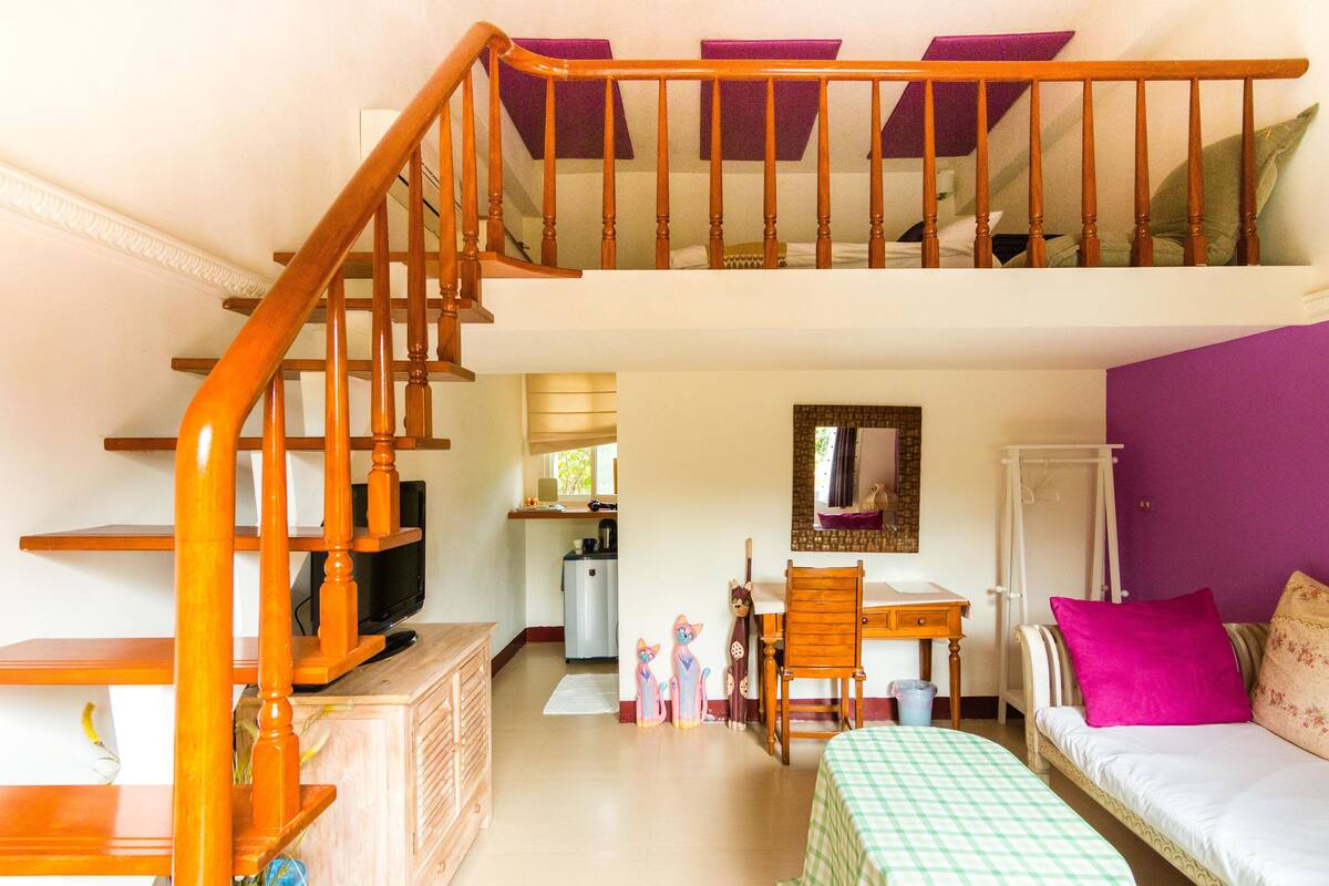 A room with a wooden staircase, a bed with a checkered bedspread, a desk with a chair, a small kitchenette, and a purple wall.