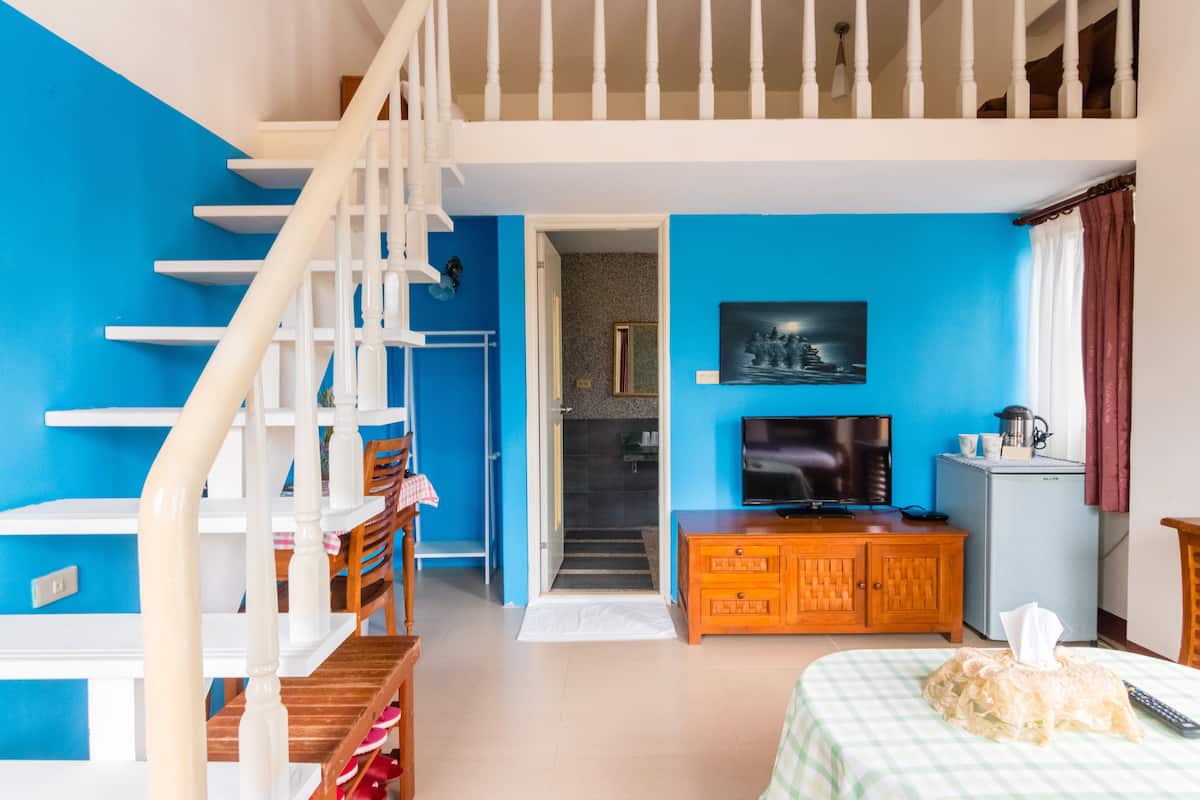 A room with a blue wall, a wooden staircase, a television, a refrigerator, and a bed with a checkered bedspread.