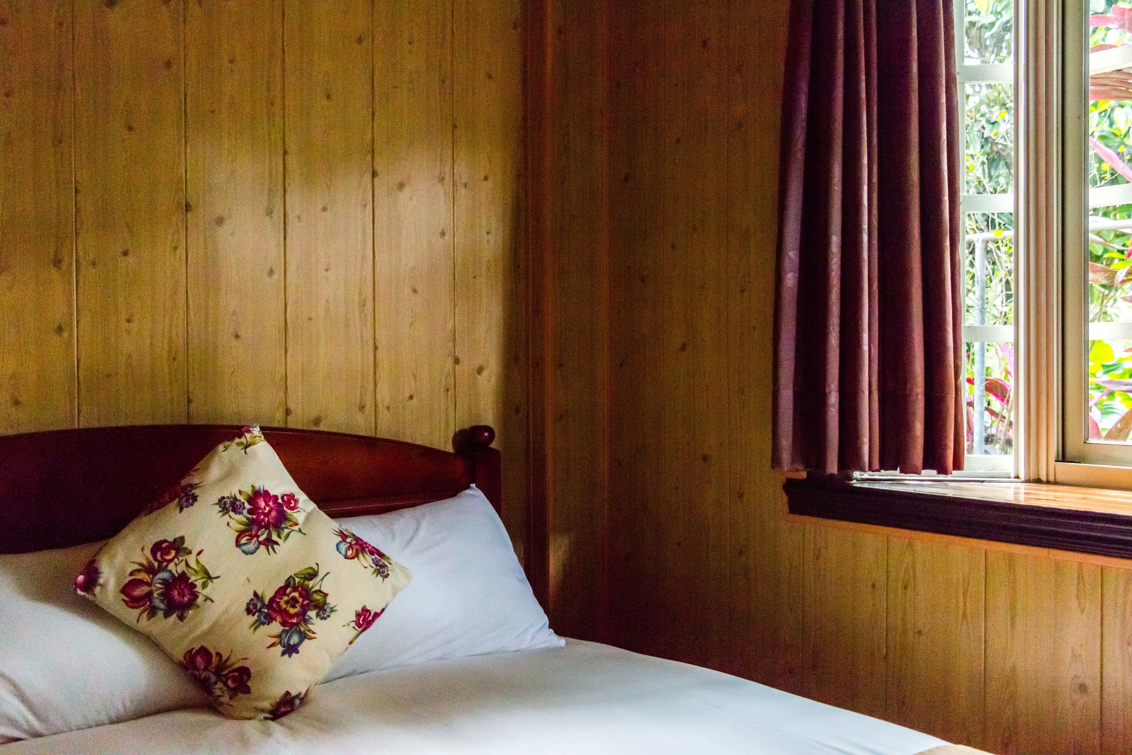 A wooden paneled room with a bed, a window with curtains, and a view of greenery outside.