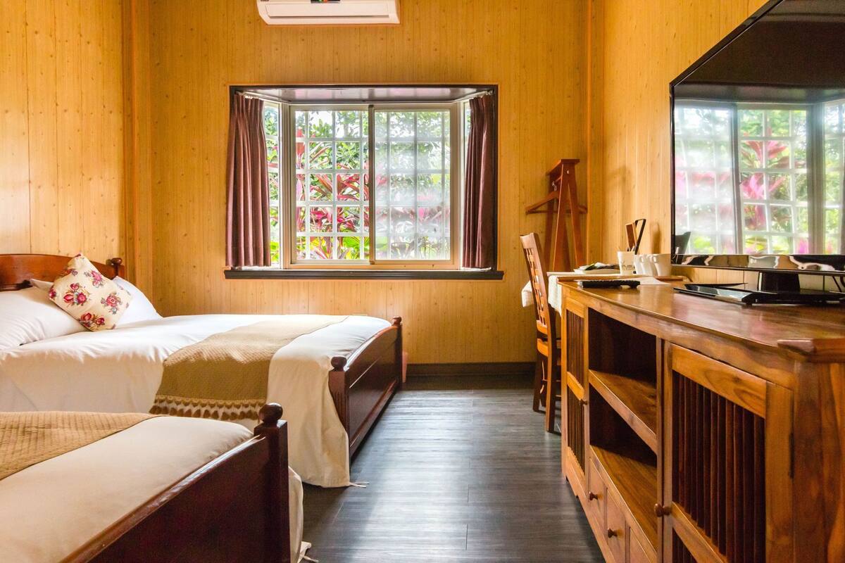 A room with two beds, a wooden desk, and a window with a view of greenery.