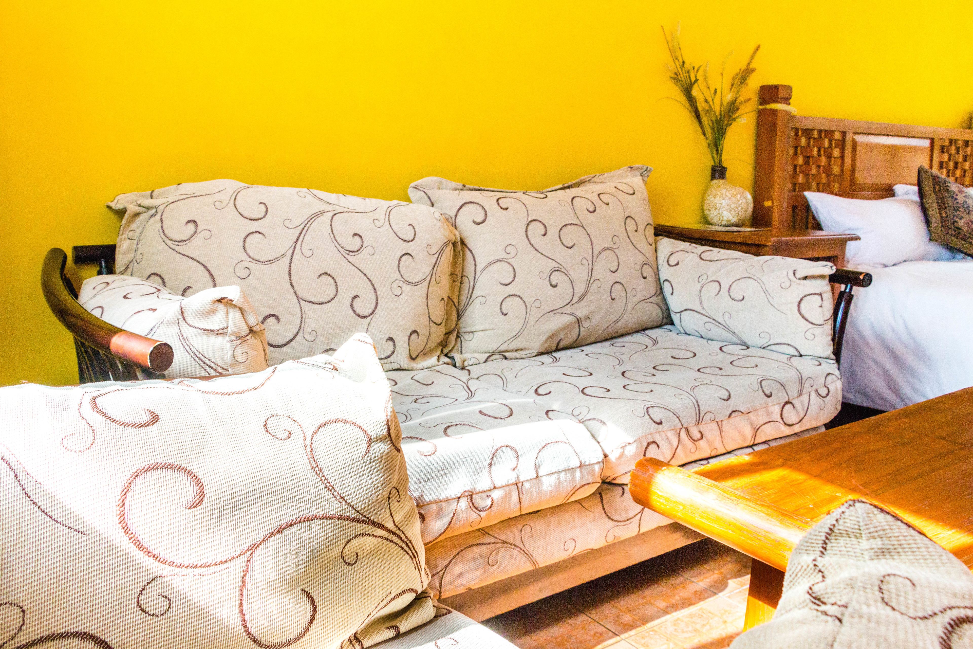 A room with a sofa set against a yellow wall, a wooden side table, and a bed with a headboard.