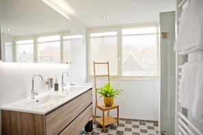 Un baño moderno con lavamanos doble, una silla de madera con planta en maceta y suelo a cuadros.